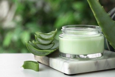 Jar with cream and cut aloe leaves on white table against blurred green background, closeup