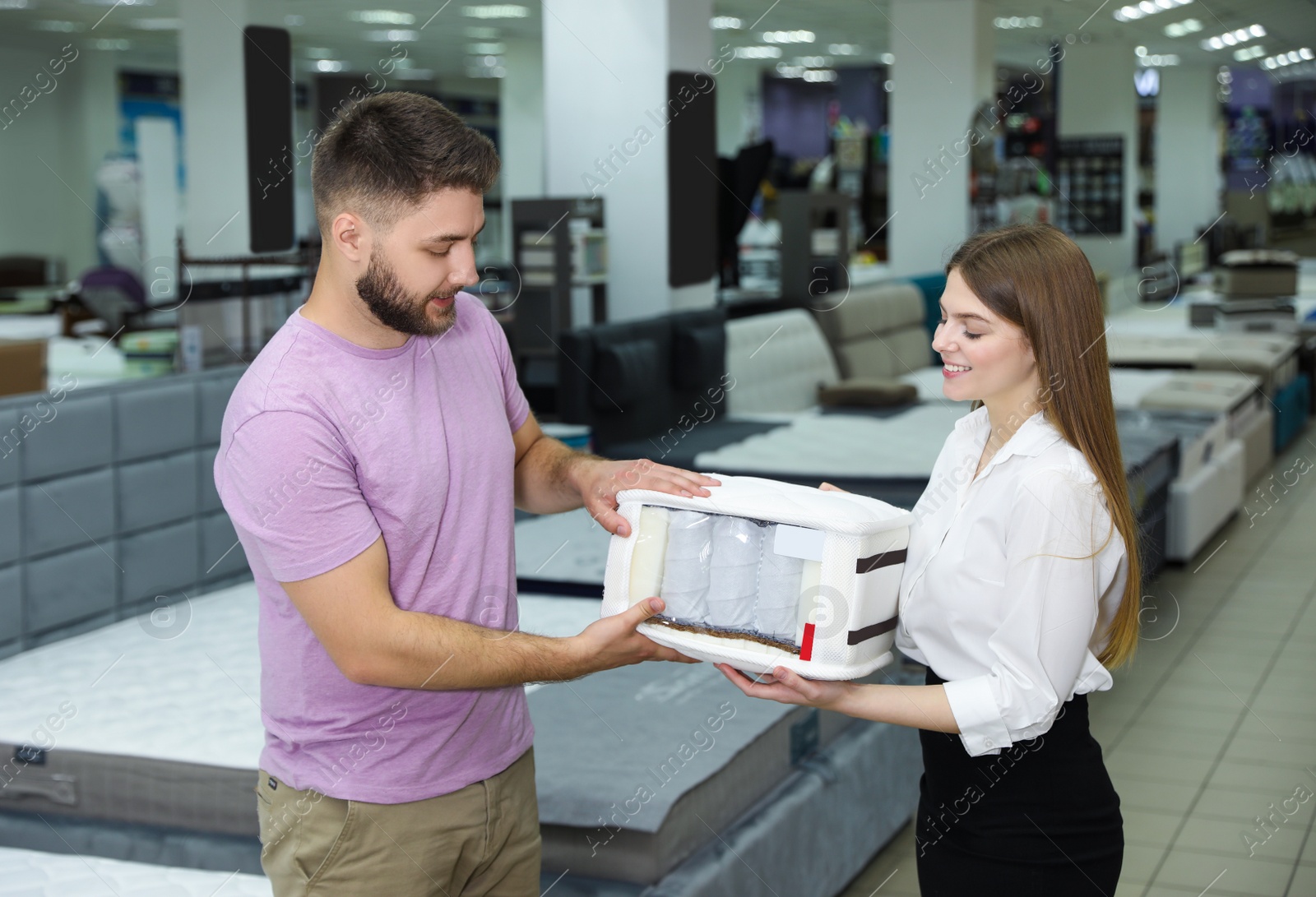 Photo of Young saleswoman showing sample of modern orthopedic mattress to customer in store