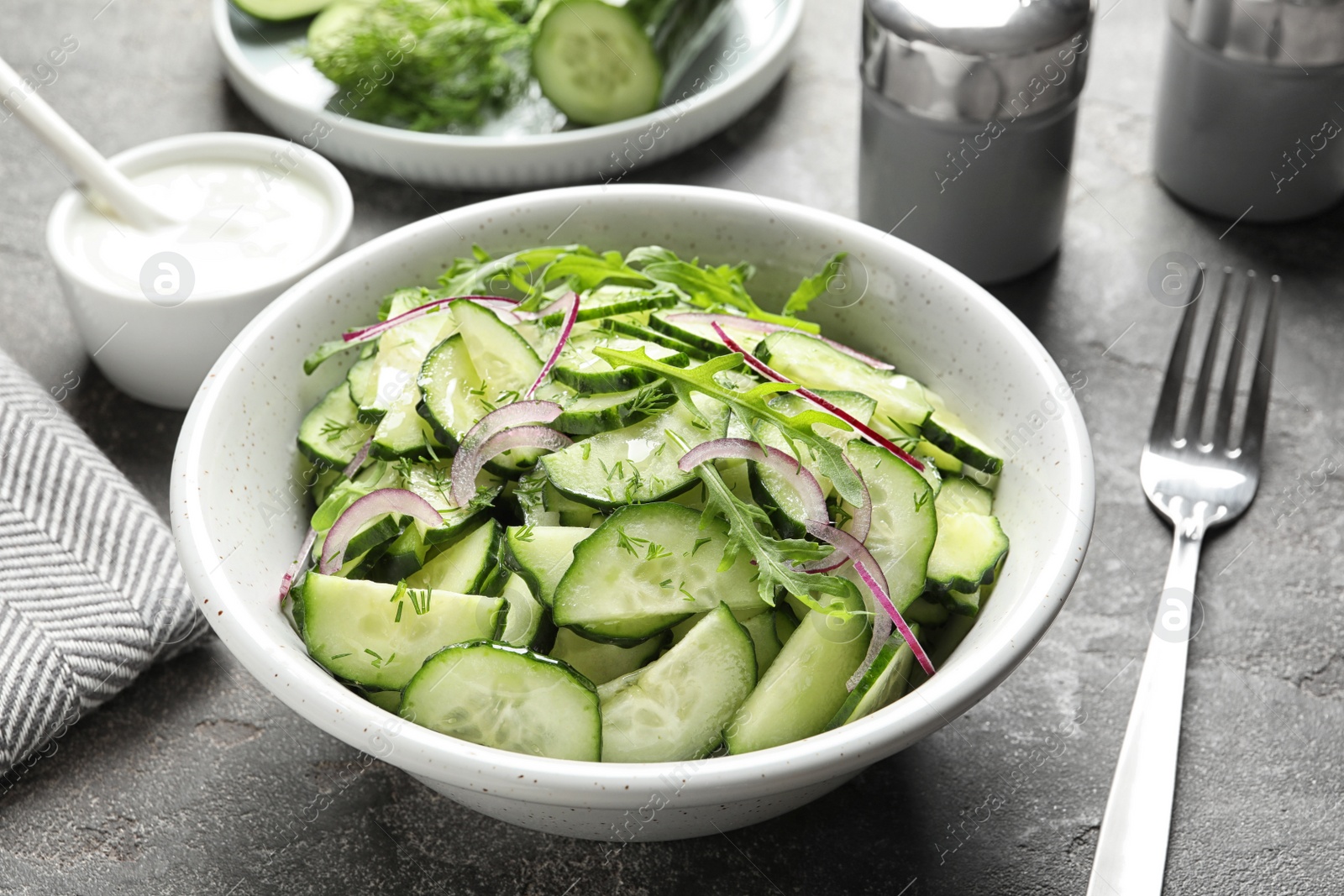 Photo of Delicious cucumber salad with onion and arugula in bowl served on grey table