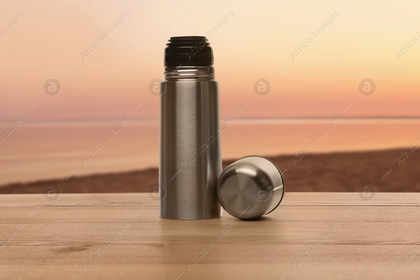 Image of Thermo bottles on wooden table near sea under sunset sky
