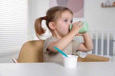 Cute little child with bottle and cup of tasty yogurt at white table indoors