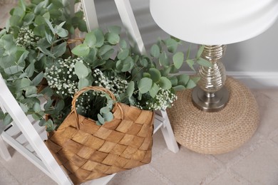 Stylish straw bag with beautiful bouquet and lamp indoors