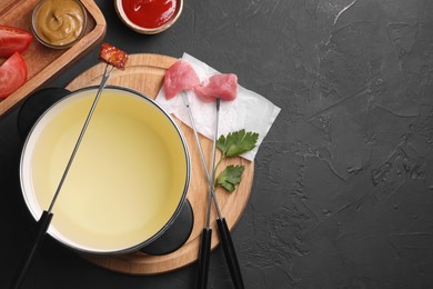 Photo of Fondue pot, forks with meat pieces and other products on black textured table, flat lay. Space for text