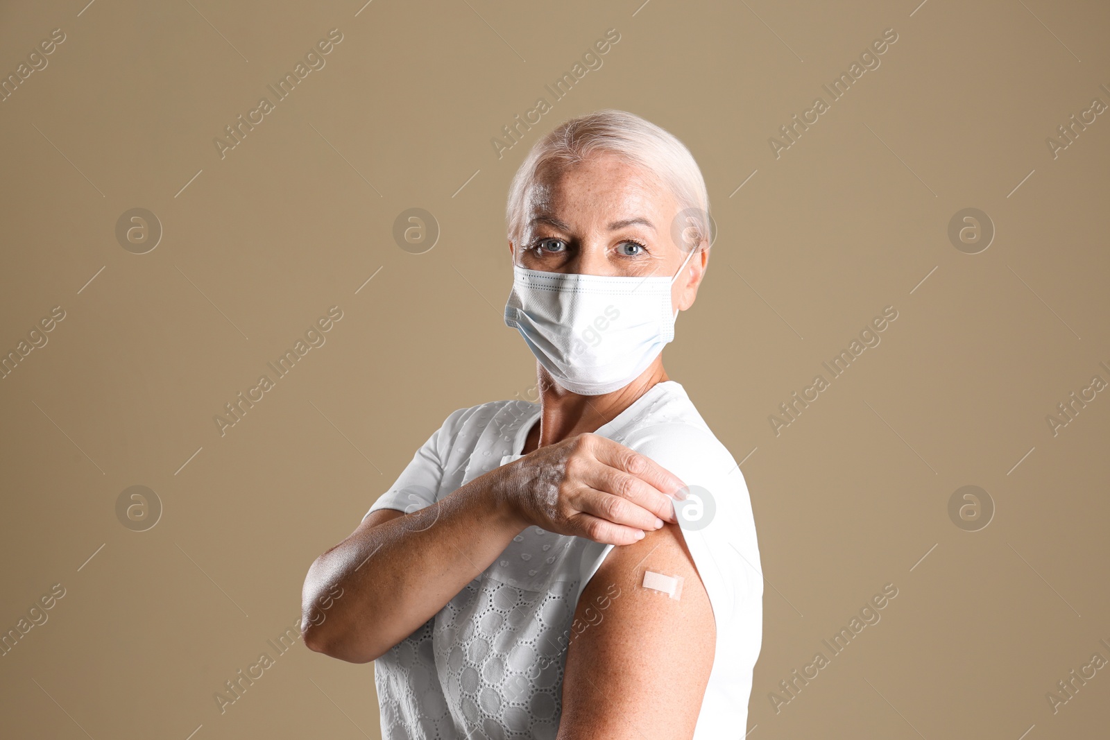 Photo of Mature woman in protective mask showing arm with bandage after vaccination on beige background