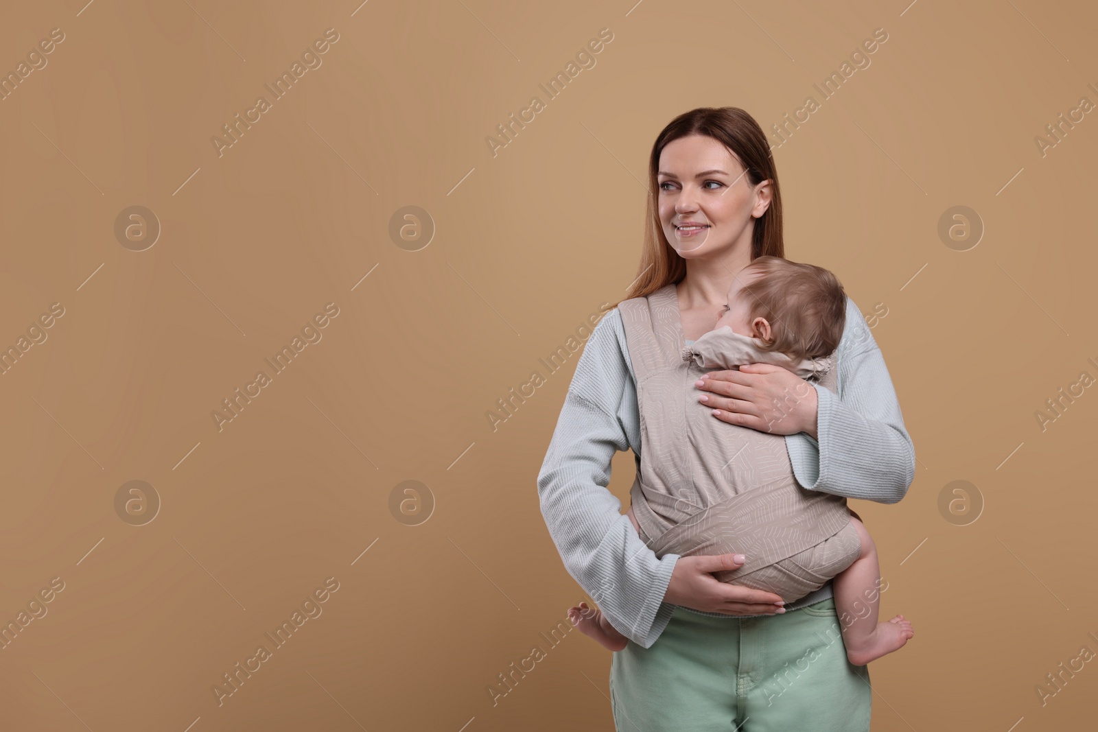 Photo of Mother holding her child in sling (baby carrier) on light brown background. Space for text