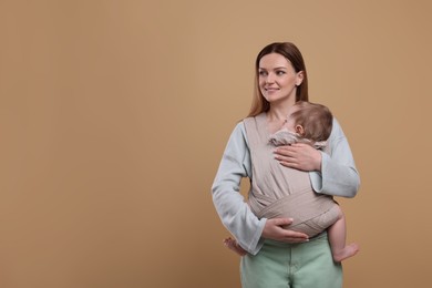 Mother holding her child in sling (baby carrier) on light brown background. Space for text