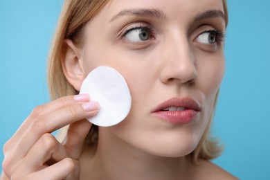Young woman cleaning face with cotton pad on light blue background, closeup