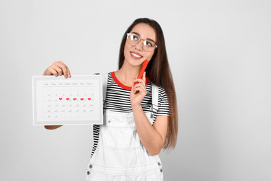 Young woman holding calendar with marked menstrual cycle days on light background