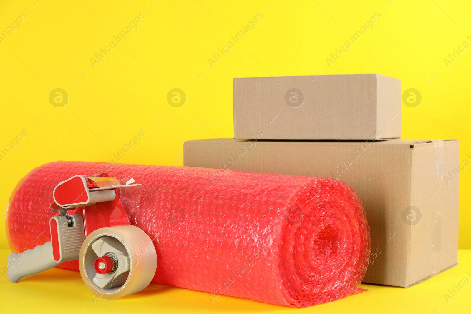 Photo of Bubble wrap roll, tape dispenser and cardboard boxes on yellow background