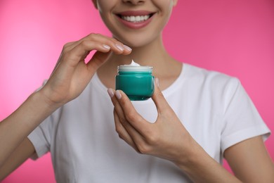 Photo of Woman holding jar of facial cream on pink background, closeup