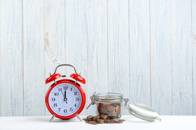 Photo of Red alarm clock, glass jar and coins on white table against wooden background. Money savings