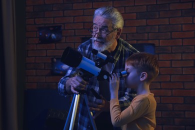 Photo of Little boy with his grandfather looking at stars through telescope in room