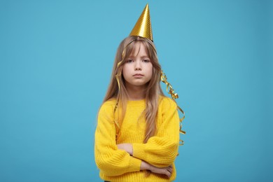 Photo of Upset little girl in party hat on light blue background