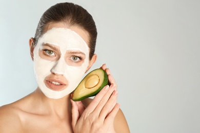 Photo of Beautiful woman holding avocado near her face with clay mask against grey background. Space for text