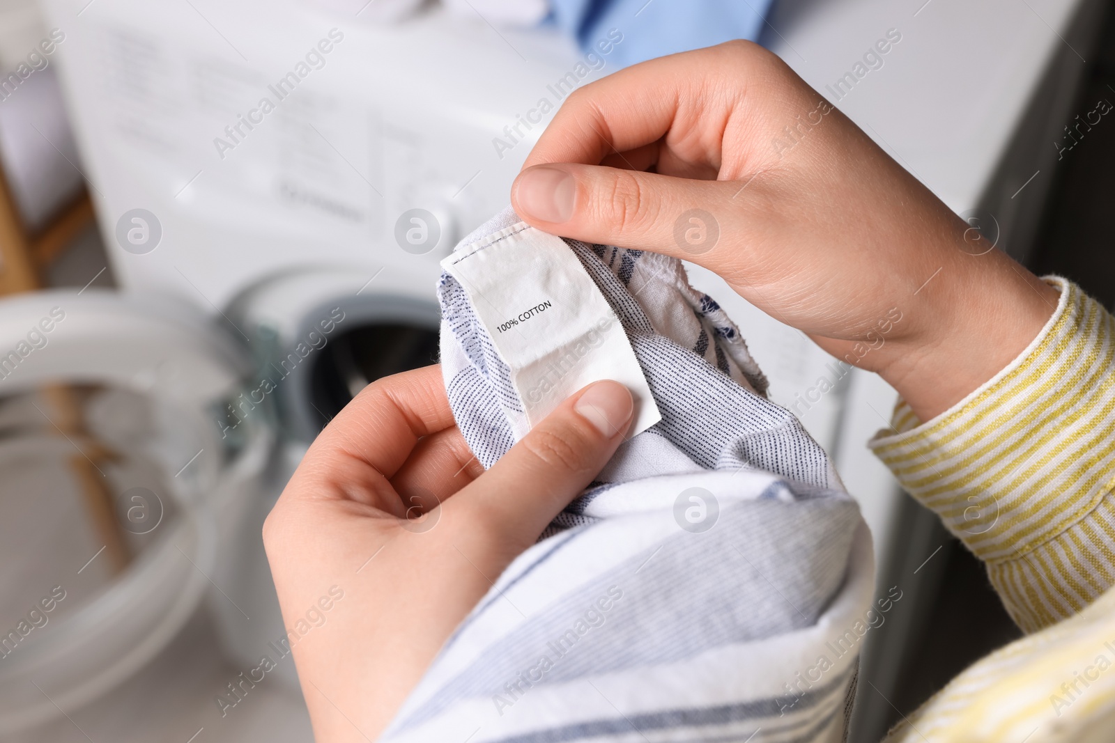 Photo of Woman reading clothing label with care symbols and material content on shirt near washing machine, closeup