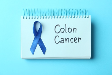 Notebook with blue ribbon and text Colon cancer on color background, top view