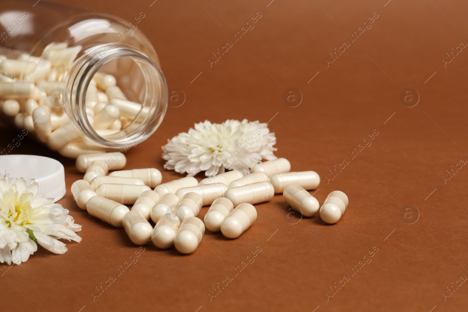 Photo of Open medicine bottle, scattered pills and flowers on brown background, space for text