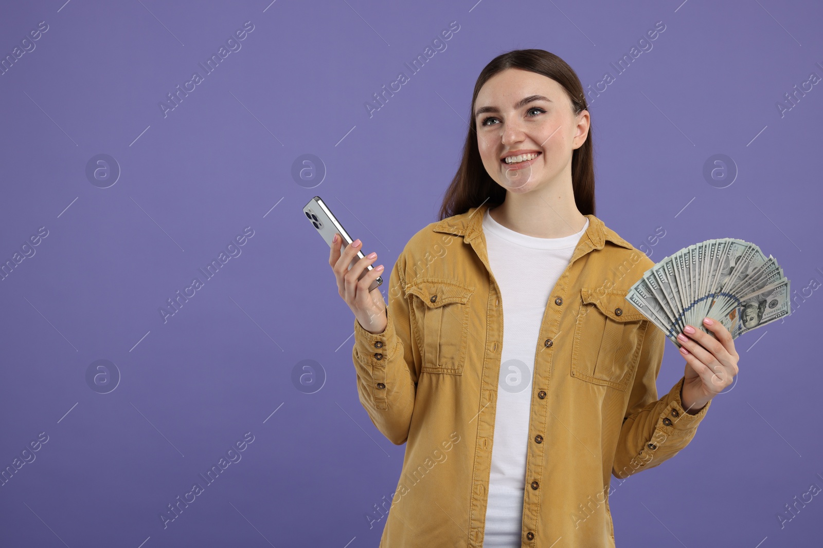 Photo of Happy woman with dollar banknotes and smartphone on purple background, space for text