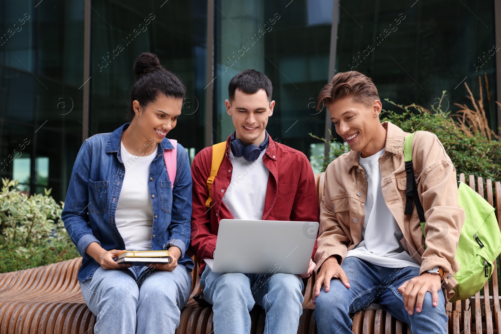 Photo of Happy young students studying with laptop on bench outdoors