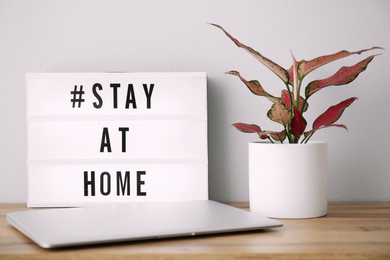 Photo of Laptop, houseplant and lightbox with hashtag STAY AT HOME on wooden table. Message to promote self-isolation during COVID‑19 pandemic