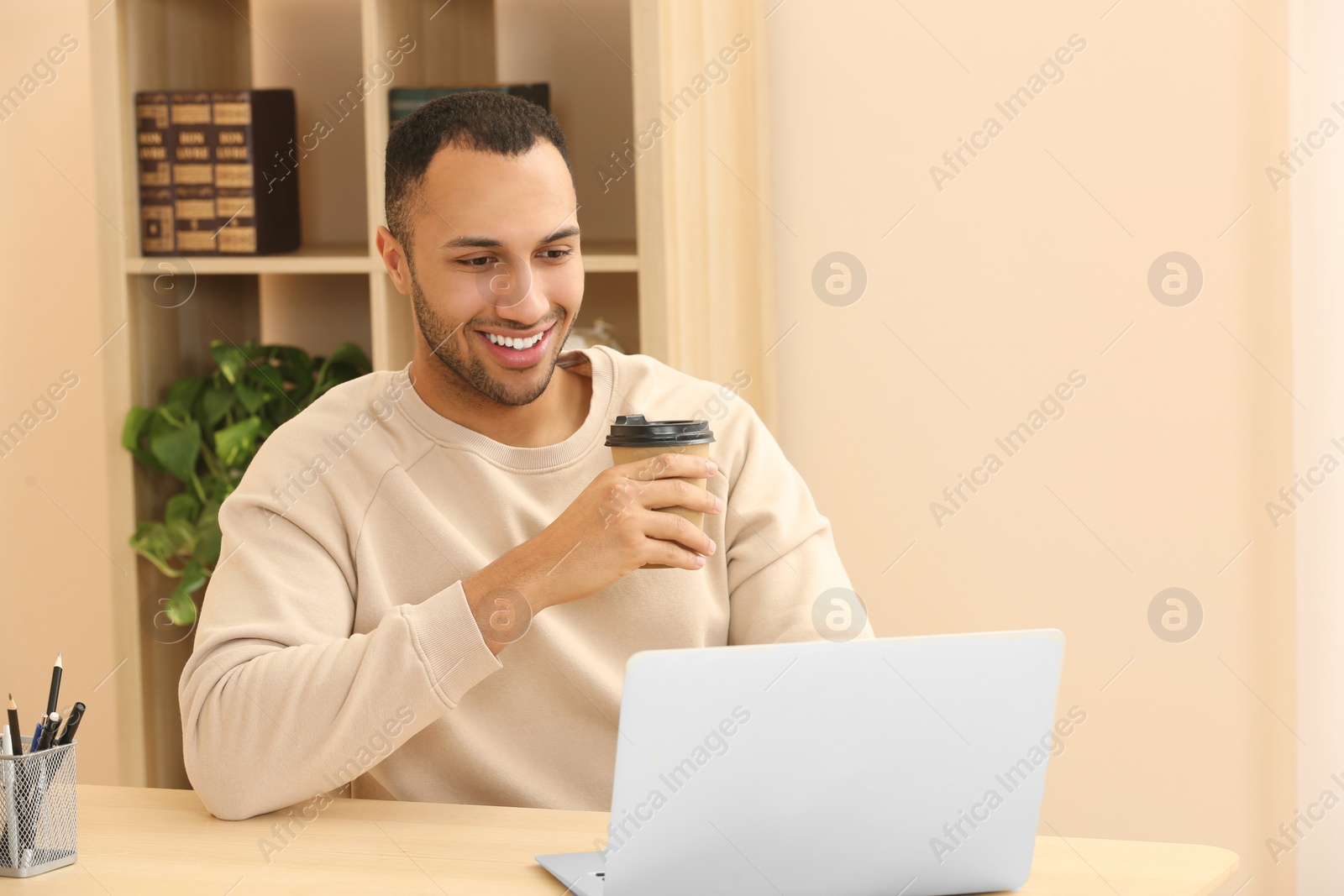 Photo of Smiling African American man with coffee spending time near laptop at wooden table in room