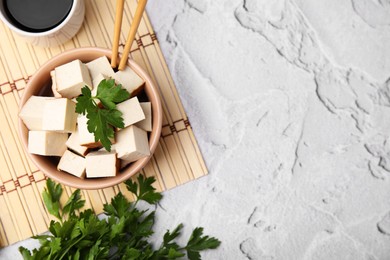 Photo of Bamboo mat with bowl of smoked tofu cubes, soy sauce and parsley on white textured table, flat lay. Space for text