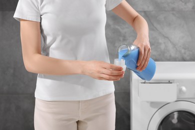 Woman pouring fabric softener from bottle into cap near washing machine indoors, closeup