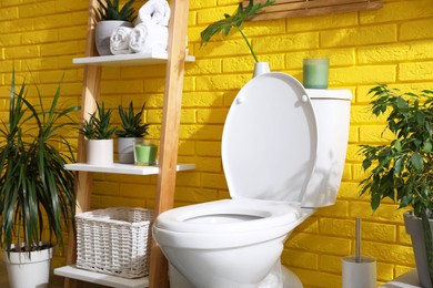 Photo of Stylish bathroom with toilet bowl and green plants near yellow brick wall. Interior design