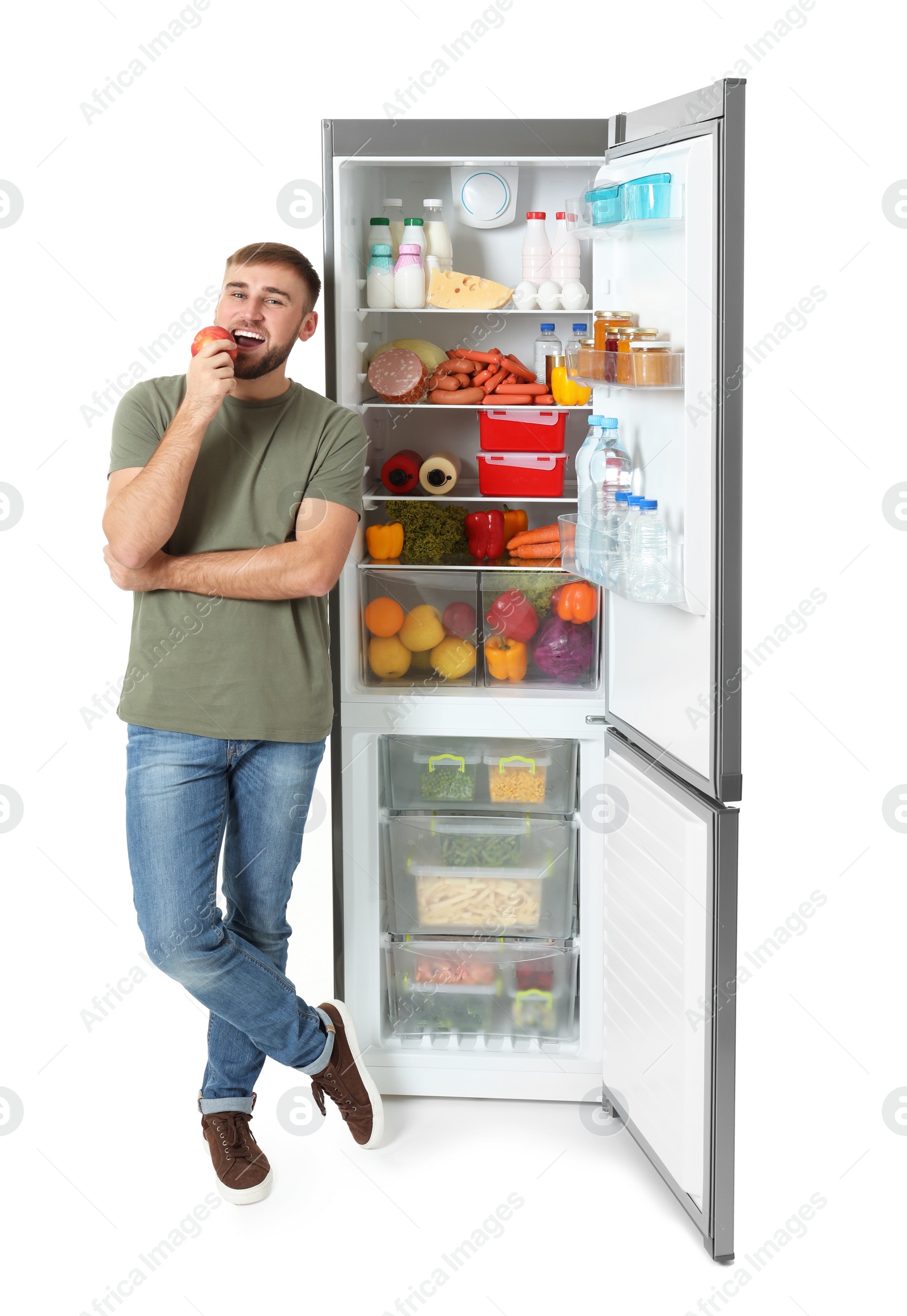 Photo of Young man eating apple near open refrigerator on white background