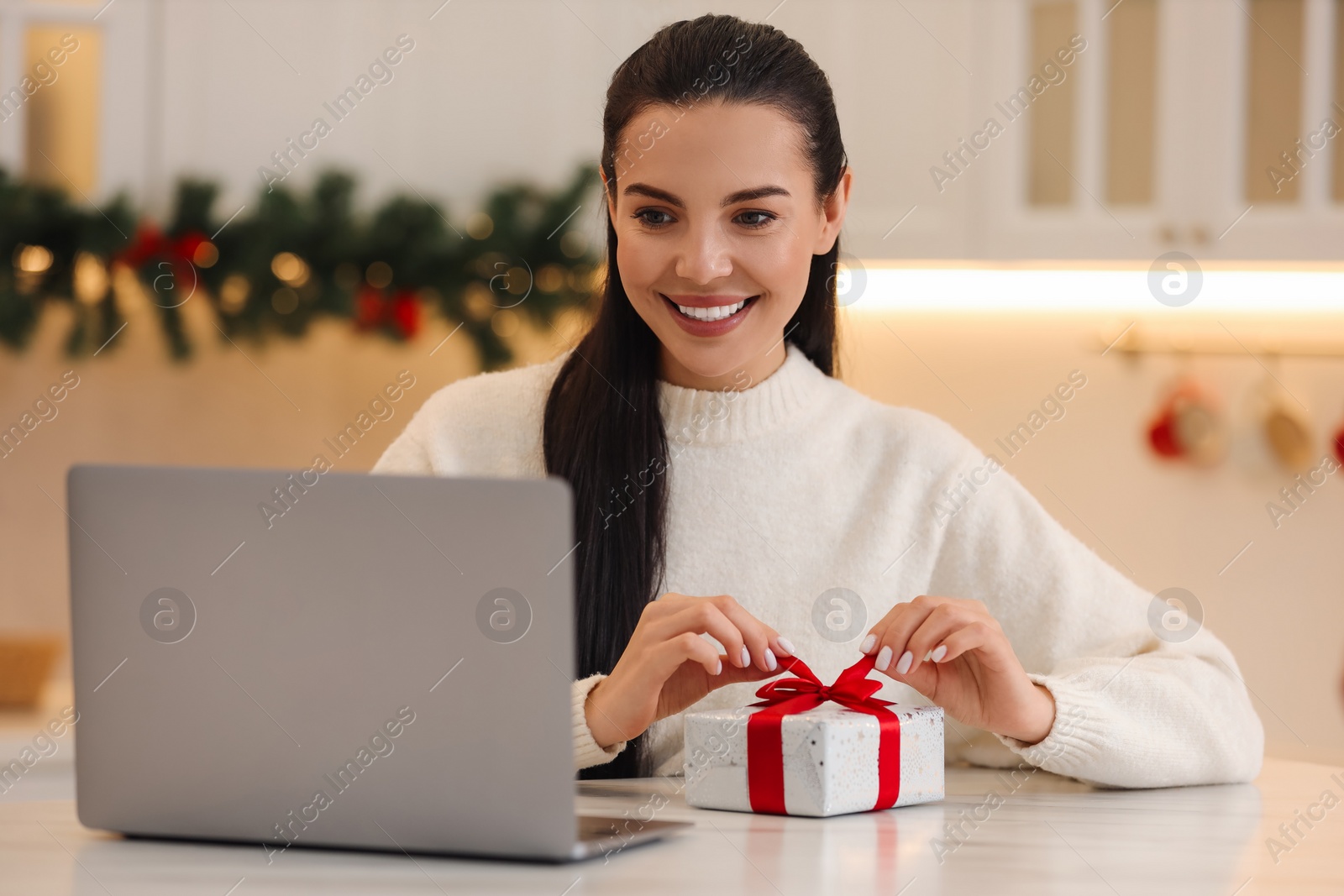 Photo of Celebrating Christmas online with exchanged by mail presents. Smiling woman opening gift box during video call on laptop at home