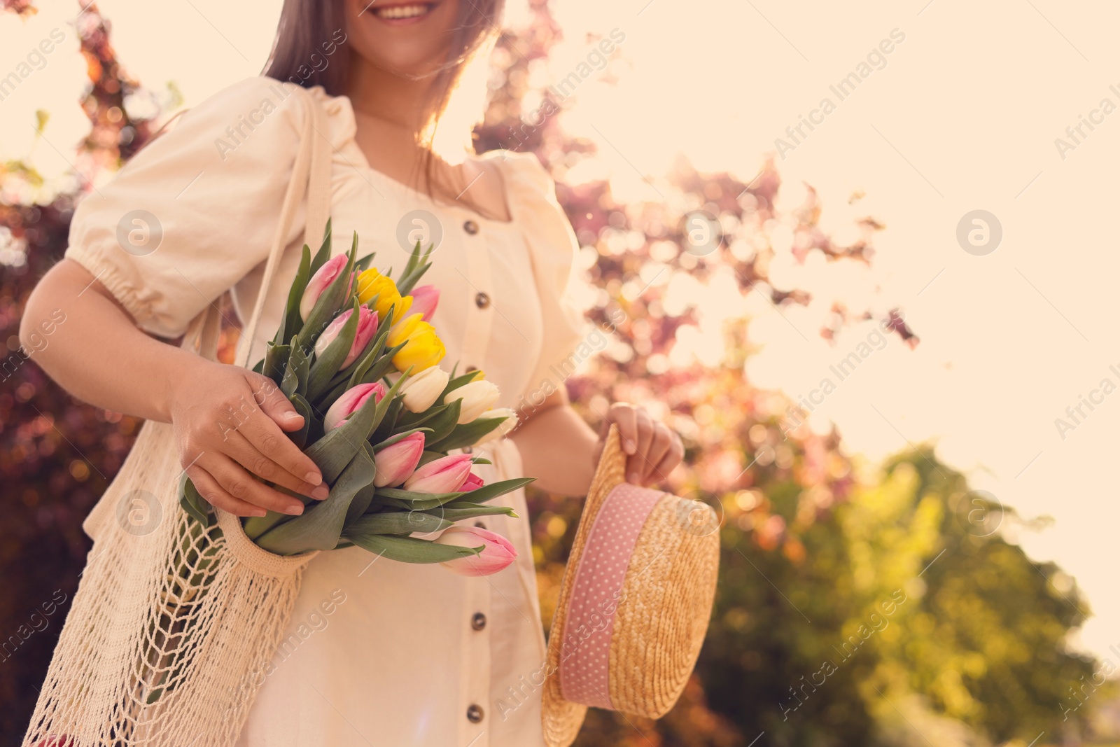 Photo of Young woman with bouquet of tulips and hat in park on sunny day, closeup. Space for text
