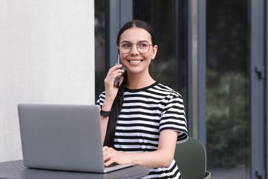 Happy young woman using modern laptop and talking on smartphone at table outdoors