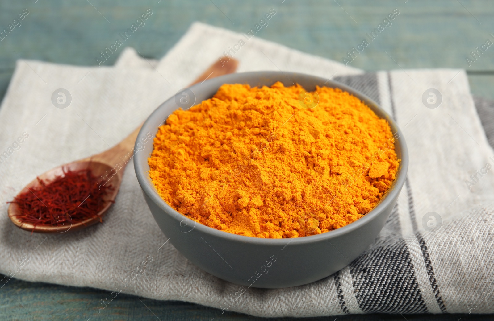 Photo of Bowl of saffron powder and spoon with dried flower stigmas on light blue wooden table