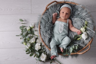 Photo of Adorable newborn baby lying in basket with faux fur and flowers on floor, top view