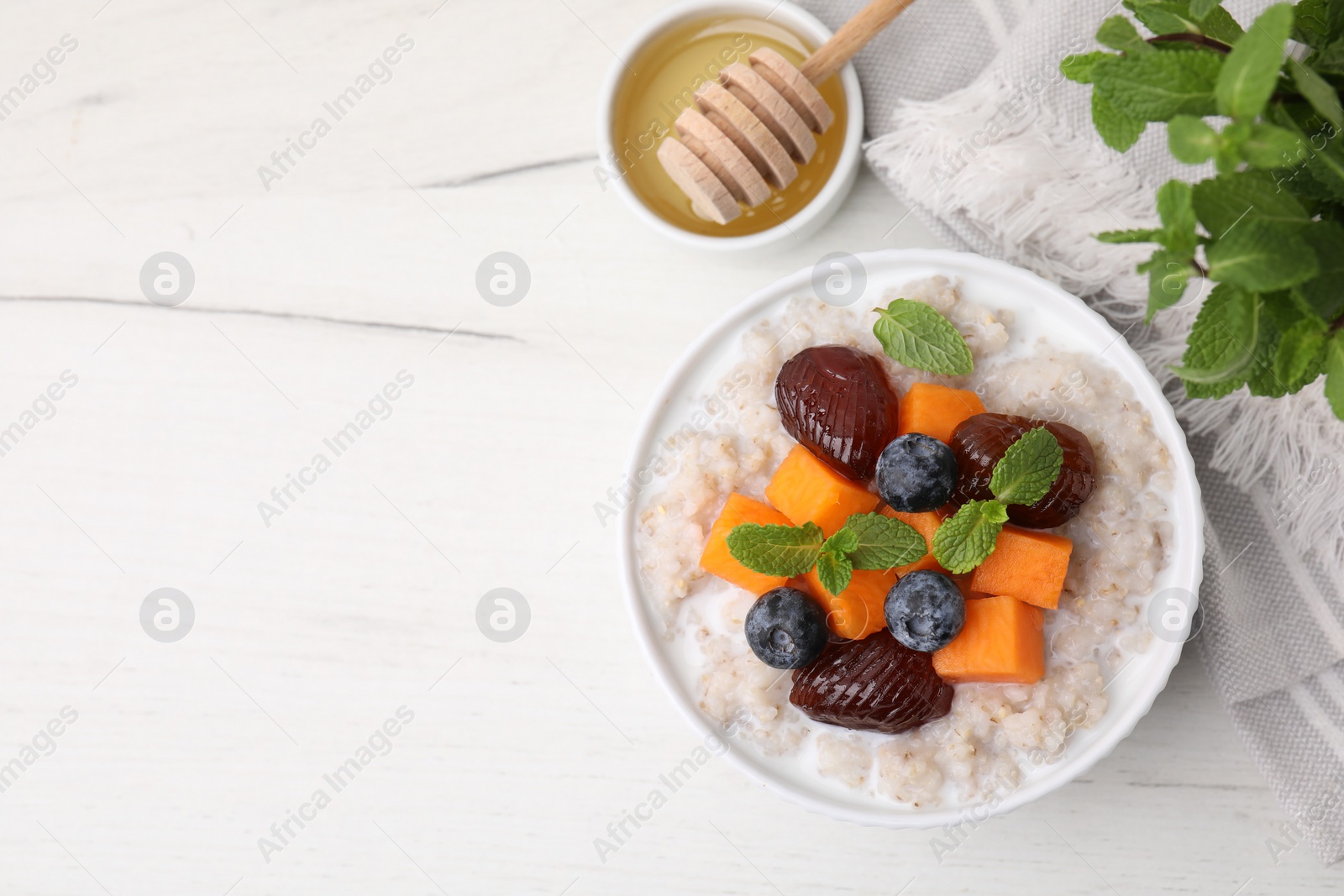 Photo of Delicious barley porridge with blueberries, pumpkin, dates and mint served with honey on white table, flat lay. Space for text