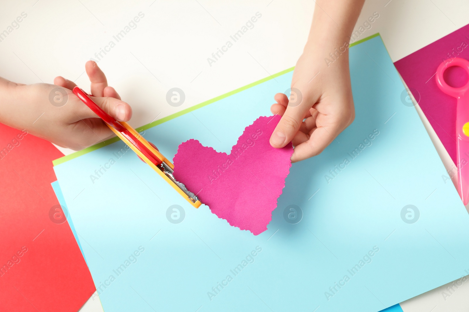 Photo of Child cutting out paper heart with craft scissors at table, top view