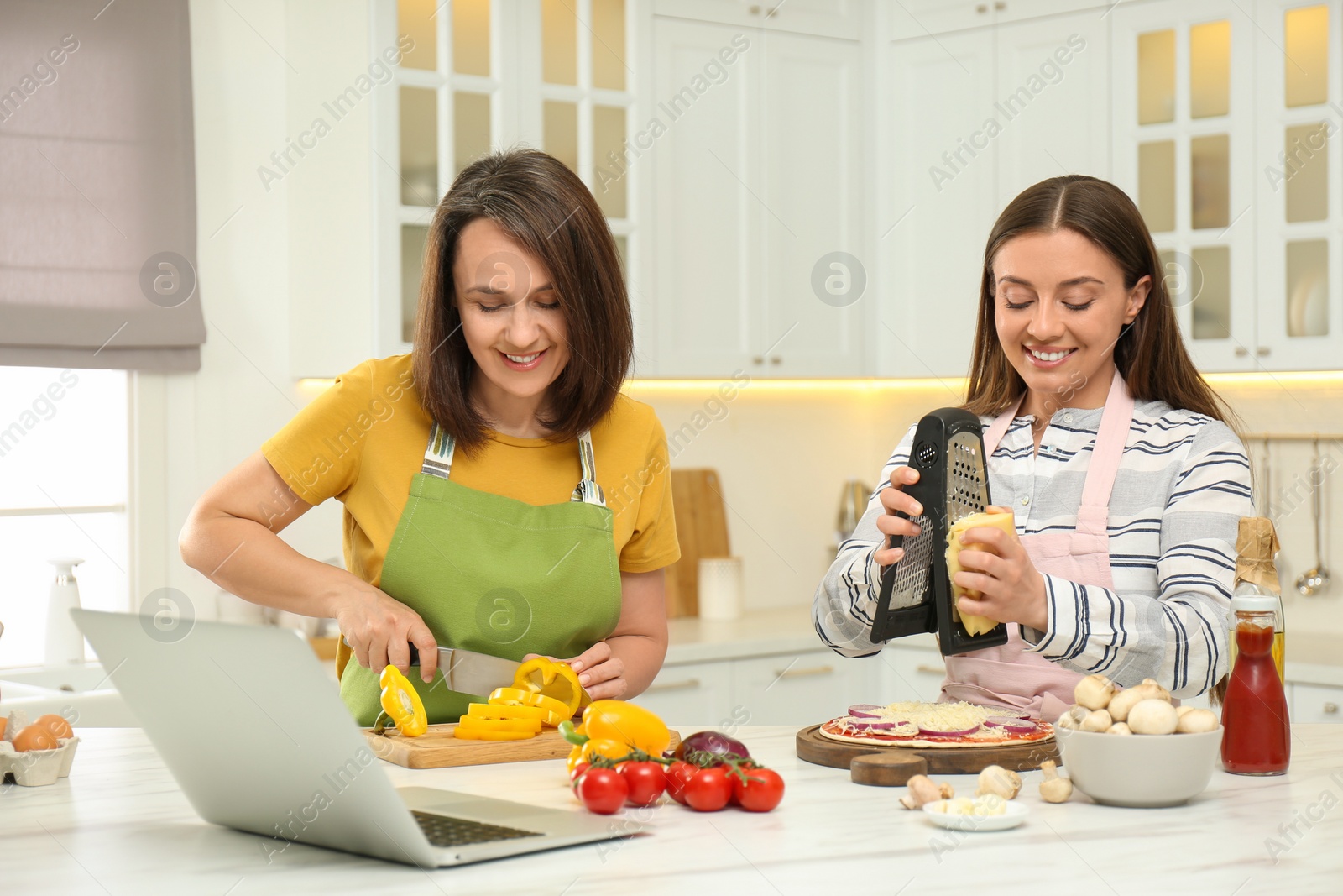 Photo of Mother with adult daughter making dinner while watching online cooking course via laptop in kitchen