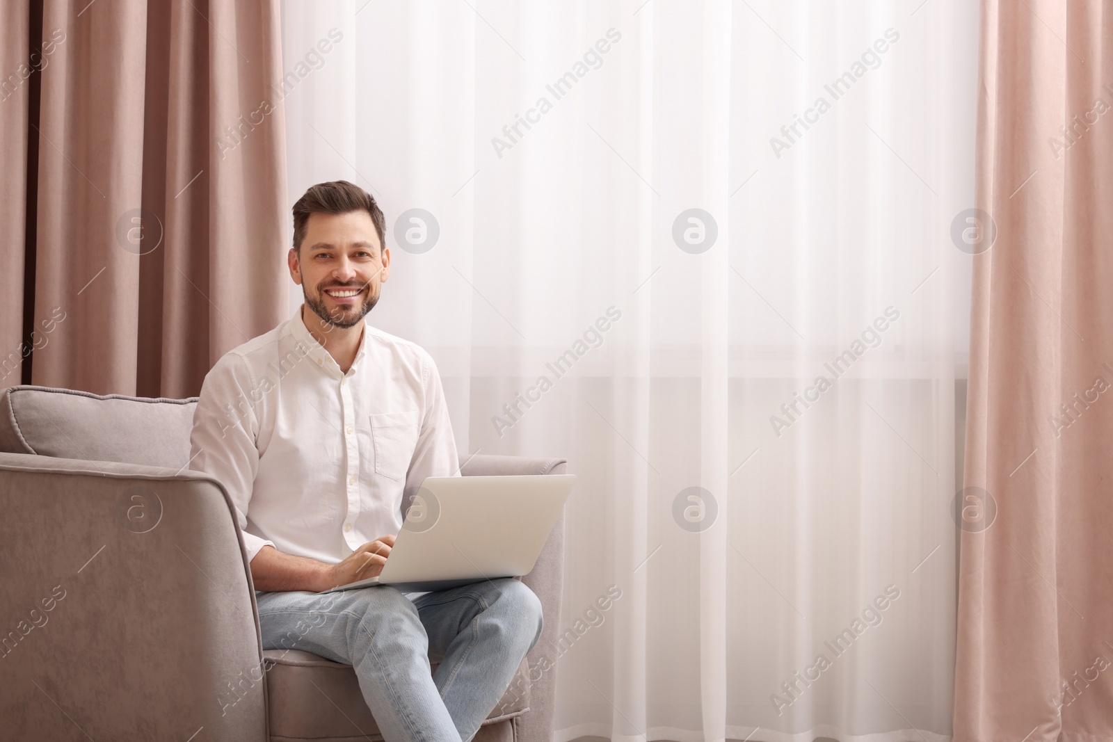 Photo of Smiling man holding laptop on armchair near window with beautiful curtains at home. Space for text