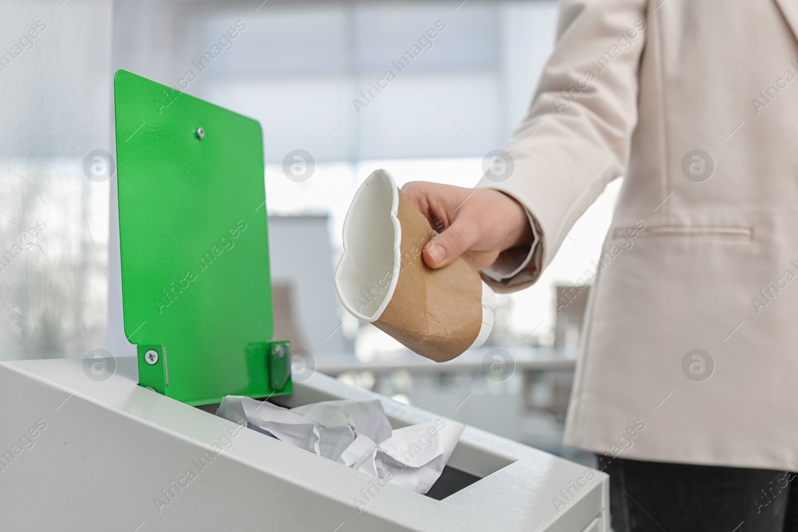 Photo of Woman putting used paper cup into trash bin in modern office, closeup. Waste recycling