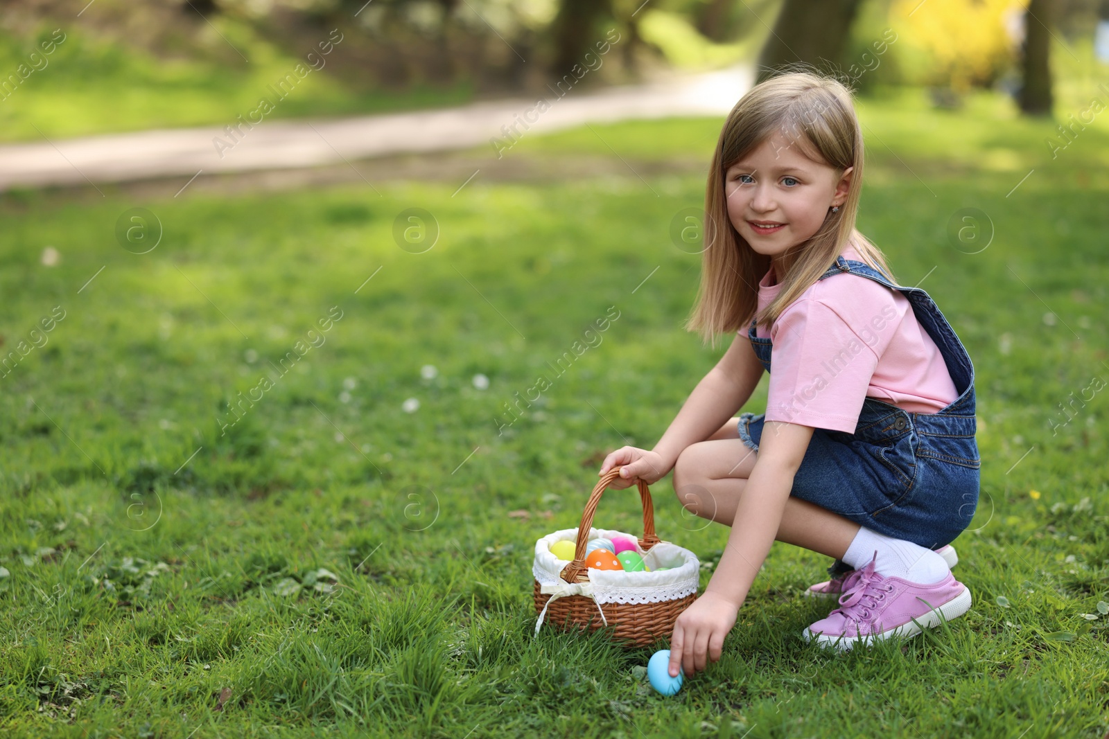 Photo of Easter celebration. Cute little girl with bunny ears hunting eggs outdoors, space for text