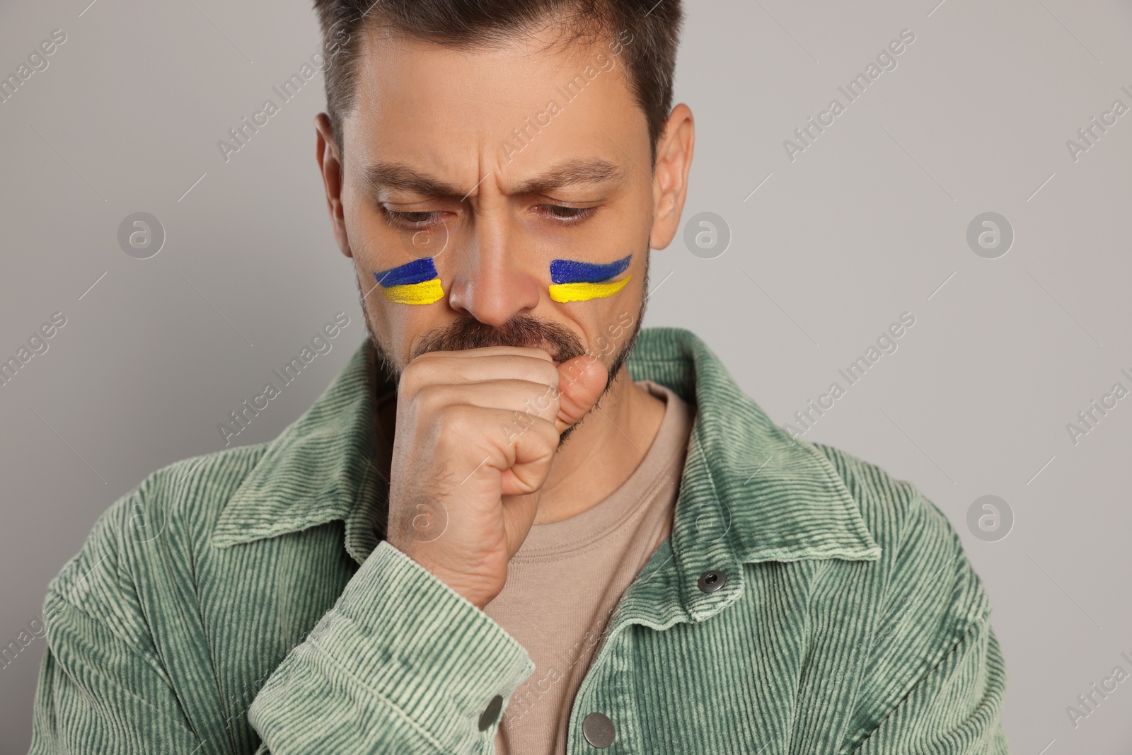 Photo of Sad man with drawings of Ukrainian flag on face against light grey background, closeup