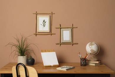 Photo of Potted houseplants, globe and stationery on wooden table near brown wall with stylish bamboo frames