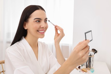 Beautiful young woman applying eyeshadow with brush at dressing table indoors