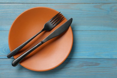 Photo of Orange ceramic plate with cutlery on light blue wooden table, top view. Space for text