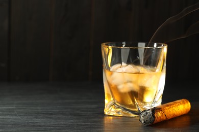 Photo of Glass of whiskey with ice cubes and smoldering cigar on black wooden table, closeup. Space for text