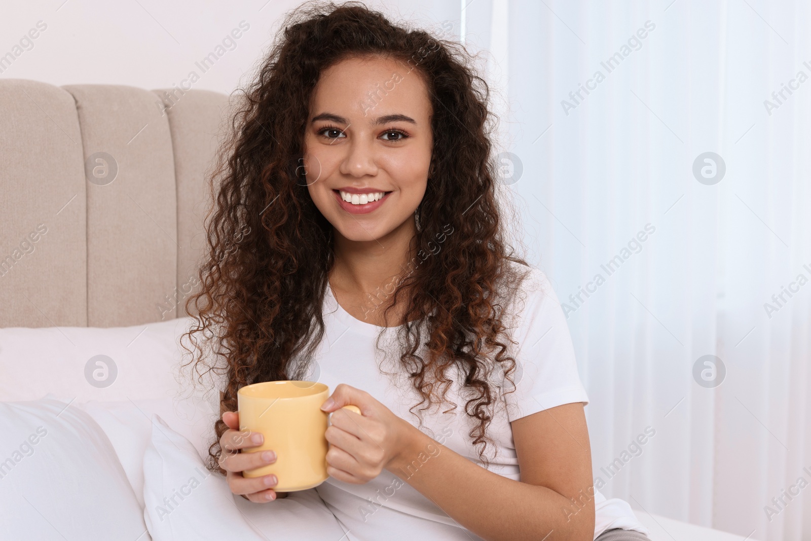 Photo of Happy African American woman with cup of drink in bed at home