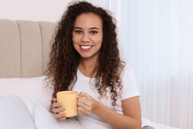 Happy African American woman with cup of drink in bed at home