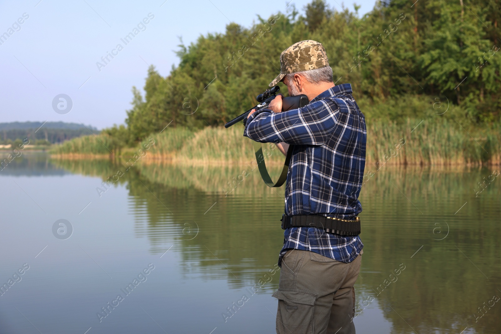 Photo of Man aiming with hunting rifle near lake outdoors. Space for text