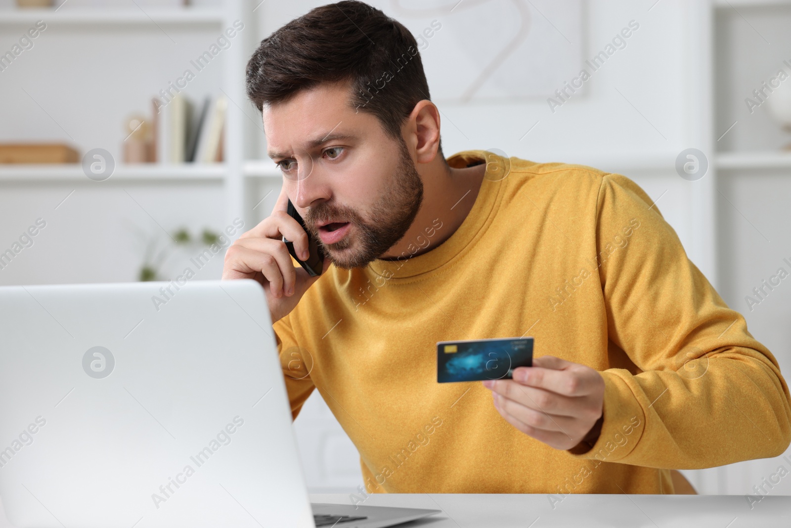 Photo of Upset man with credit card talking on smartphone near laptop at home. Be careful - fraud