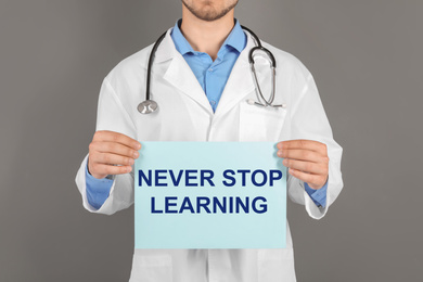 Professional doctor holding sheet of paper with phrase NEVER STOP LEARNING on grey background, closeup